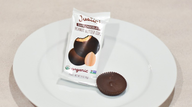 A chocolate peanut butter cup on a white plate next to a Justin's dark chocolate peanut butter cup wrapper