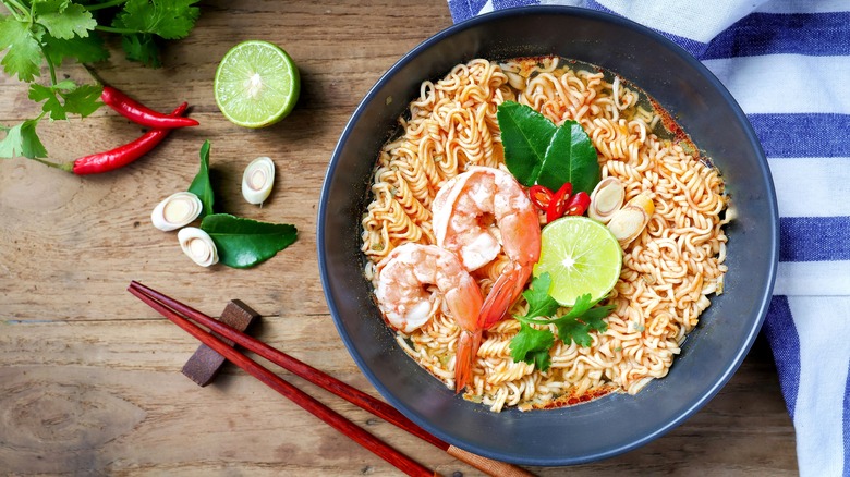 a bowl of noodles and shrimp with lemon, chilis, and chopsticks on the side