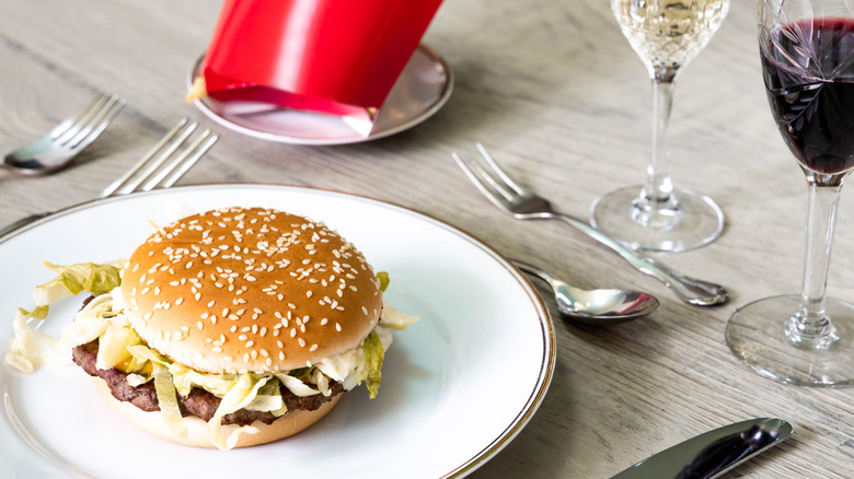 Fast food hamburger on white china with French fries carton, a glass of wine in a cut crystal glass and nice cutlery