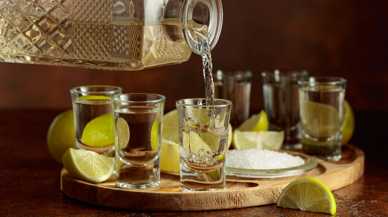 Tequila being poured into shot glasses surrounded by limes