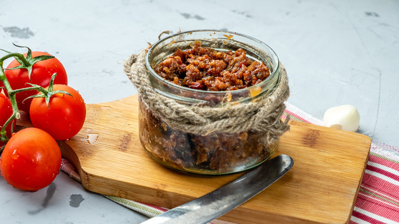 A jar of bacon jam on a cutting board beside tomatoes on the vine.