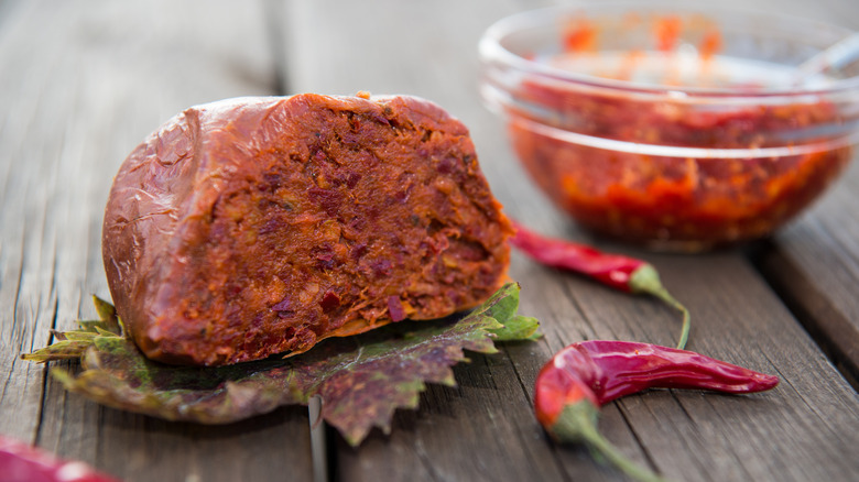 A hunk of 'nduja, spreadable pork sausage, is pictured on a wooden table with peppers.
