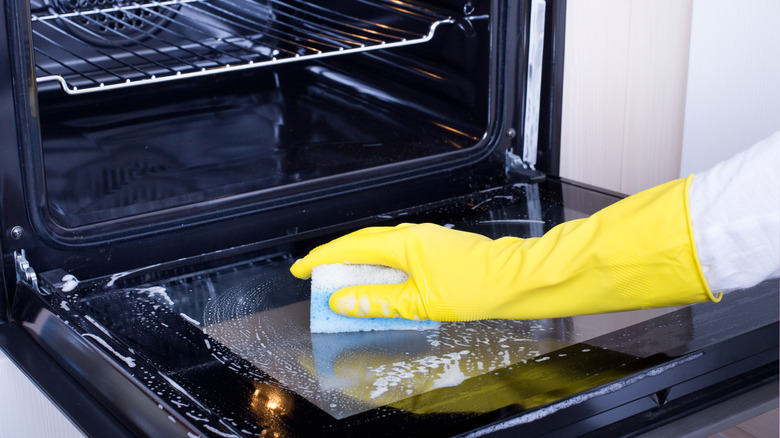 Yellow gloved hand cleaning oven glass door with soapy sponge