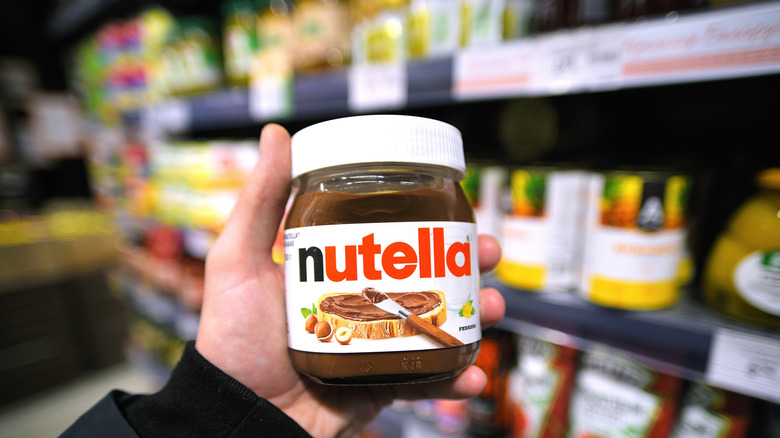 A hand holding a Nutella jar with a grocery aisle as a backdrop.