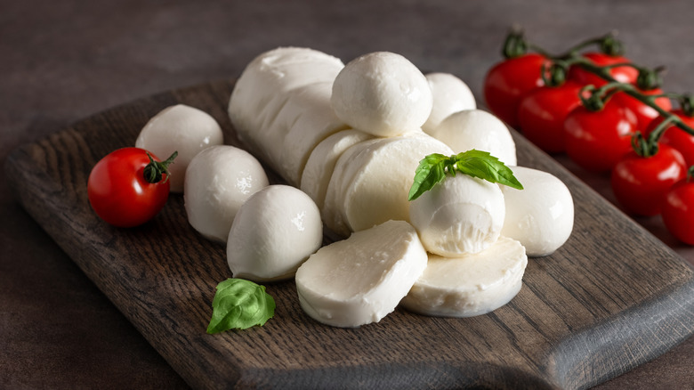 Sliced and whole fresh mozzarella on a wooden cutting board next to grape tomatoes and micro basil leaves
