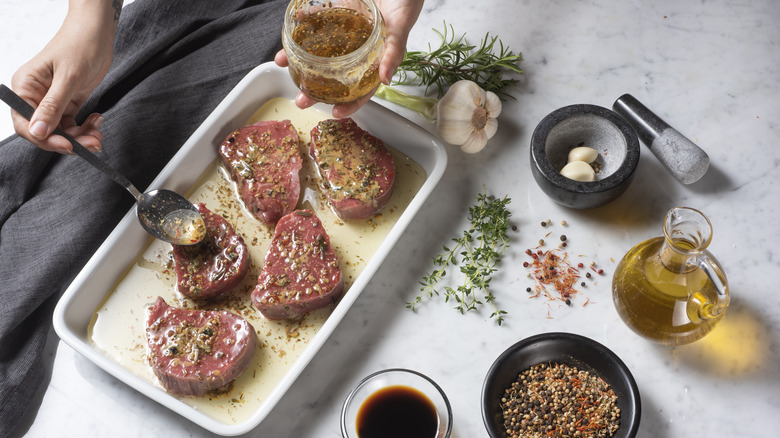 Person spooning marinade over meat in a white baking dish