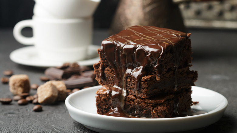 A stack of three brownies topped with glaze on a white plate in front of a stack of mugs