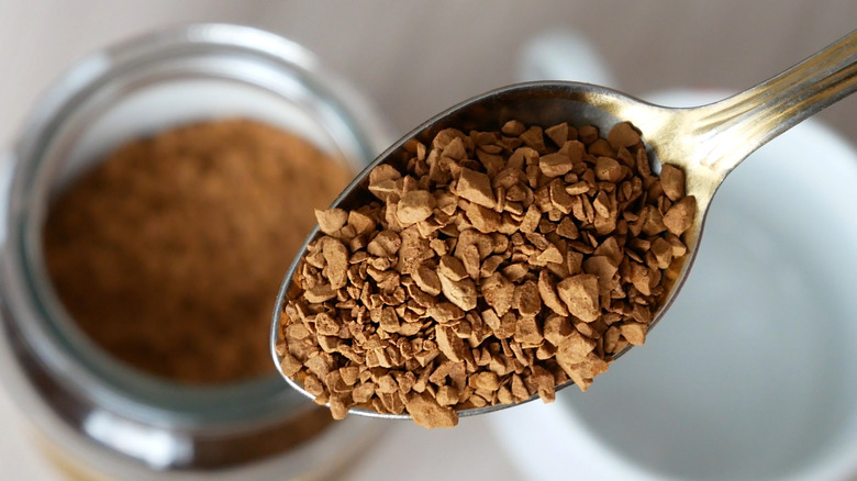 A spoonful of instant coffee granules over a white mug and glass jar