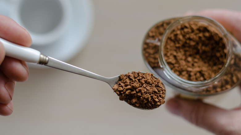 A hand holding a spoon with instant coffee granules next to a glass jar with more instant coffee granules.