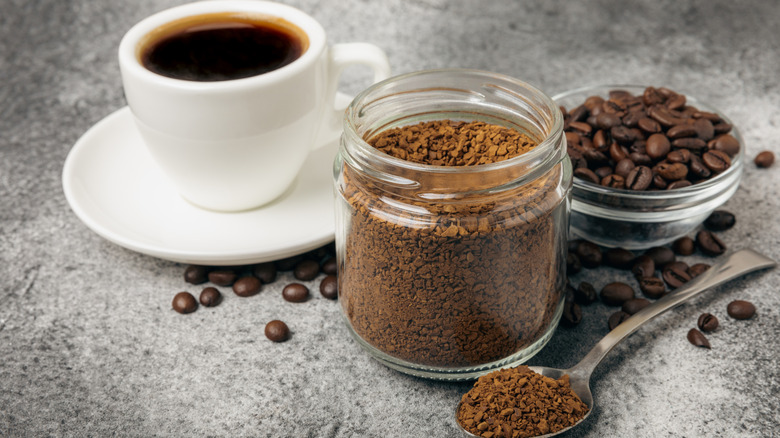 A cup of coffee on a saucer, a jar and spoon holding instant coffee granules, and a glass bowl with coffee beans