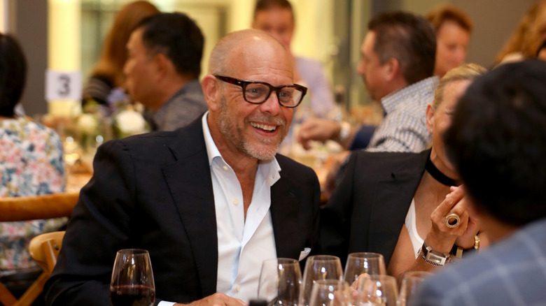 Alton Brown smiling while speaking to others at table.