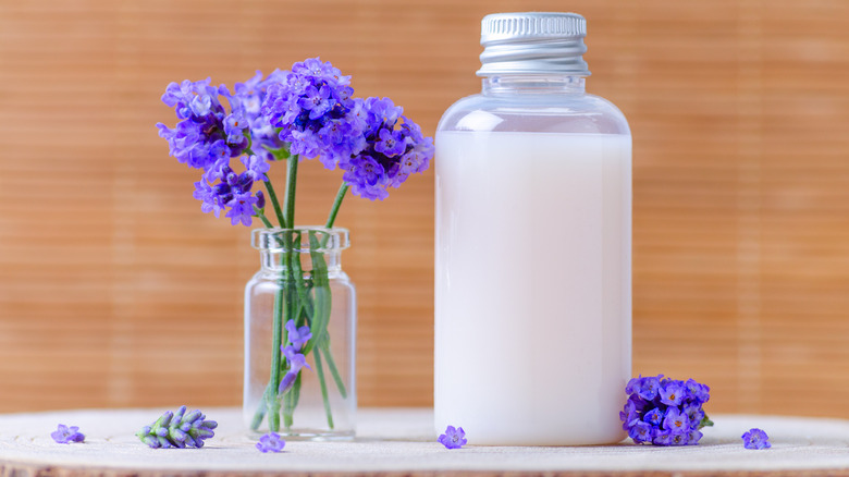 A jar of cream filled with herbs with purple flowers next to a bottle of cream.