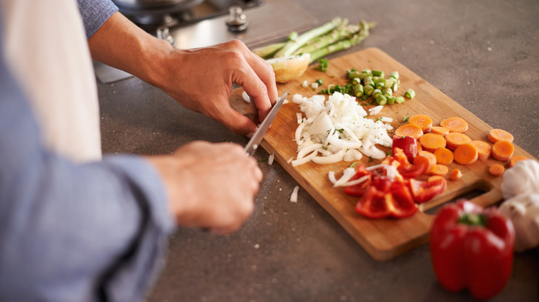 Person slicing and dicing onions, carrots, red bell pepper, asparagus, and garlic on a wooden cutting board