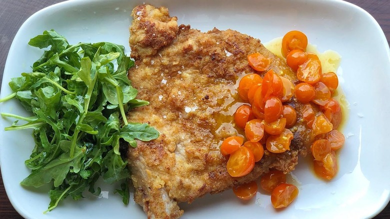 A plate with a pork chop Milanese, tomatoes, and greens from chi SPACCA