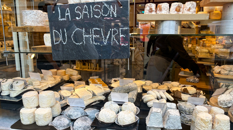 A display of goat cheeses in a storefront window with a black chalkboard sign hanging above that reads "La Saison Du Chevre"