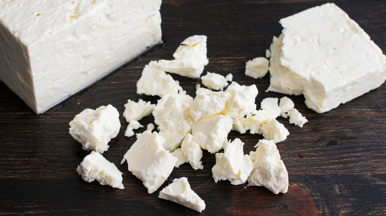 A brick of white feta cheese sits in the background with crumbled pieces of cheese in the foreground sitting on a dark wooden table