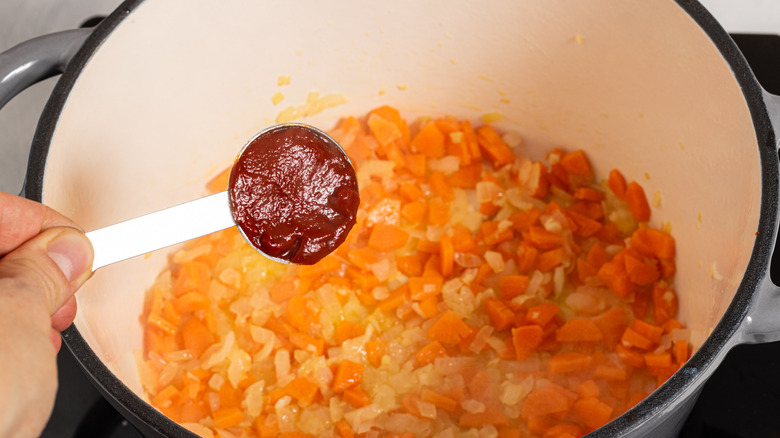 Adding tomato paste to a soup pot with chopped onions and carrots