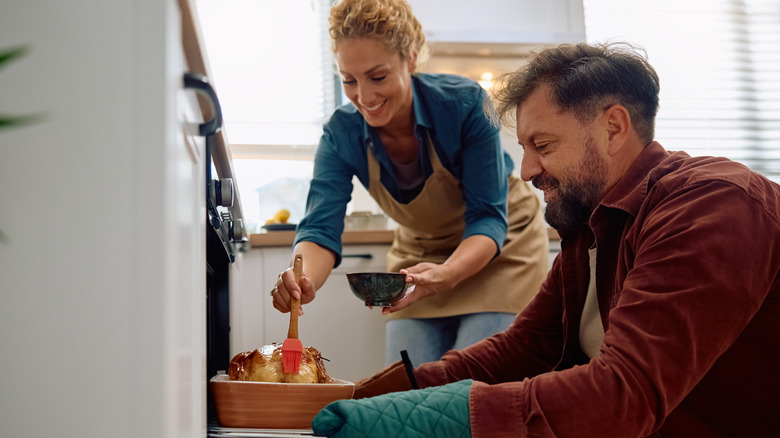 A couple is basting a turkey with the oven door open.