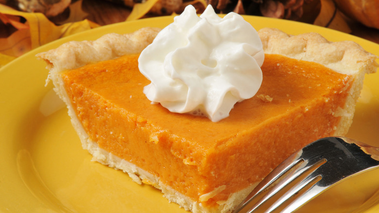 A large slice of sweet potato pie on a yellow plate, a fork next to it, topped with a dollop of whipped cream.