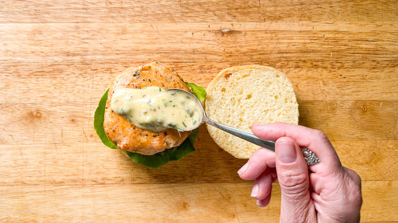 Adding remoulade sauce to broiled salmon burger on bun