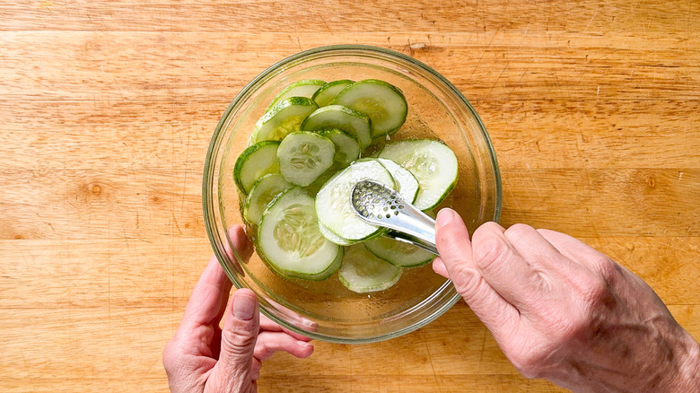 Mixing quick pickled cucumbers with tongs in glass bowl