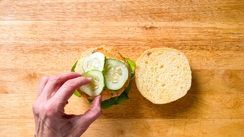 Adding pickled cucumber slices to broiled salmon burger on cutting board