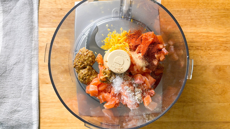 Cubed raw salmon filet with lemon zest, whole grain mustard, and spices in food processor bowl on cutting board