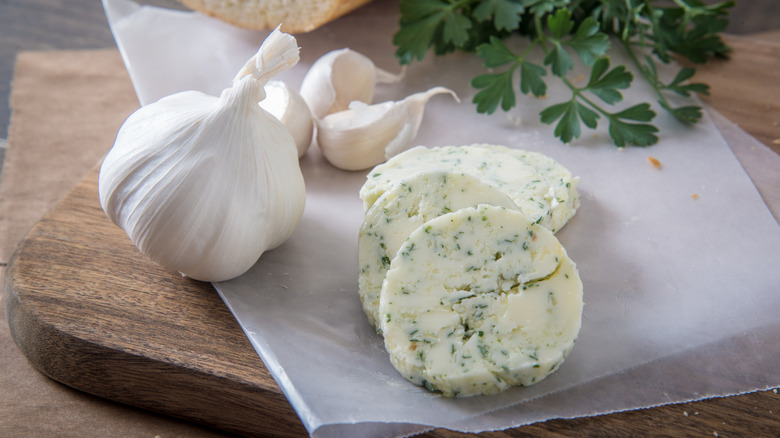 Slices of garlic butter with herbs next to a bulb of fresh garlic and herbs on parchment paper.