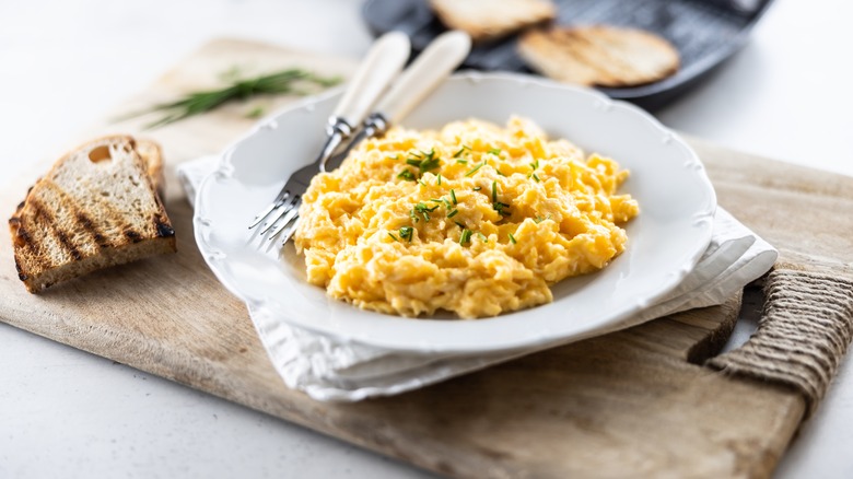 A nice plate of scrambled eggs beside toast on a cutting board.