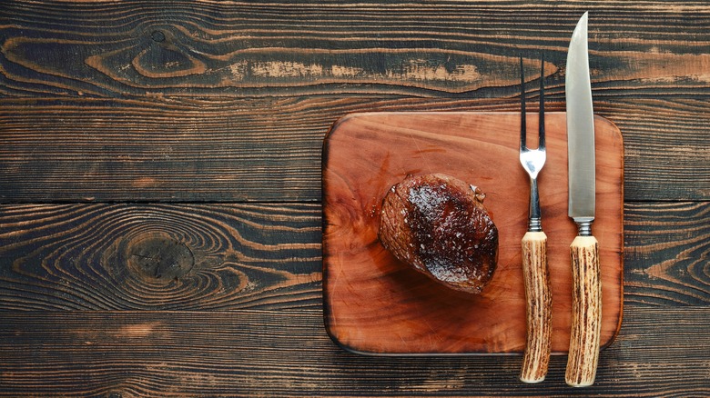 A filet, two-pronged fork, and sharp steak knife on a cutting board.