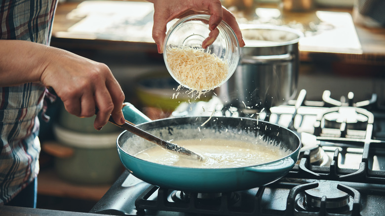 A person's hands prepare alfredo sauce in a pan on a stovetop.