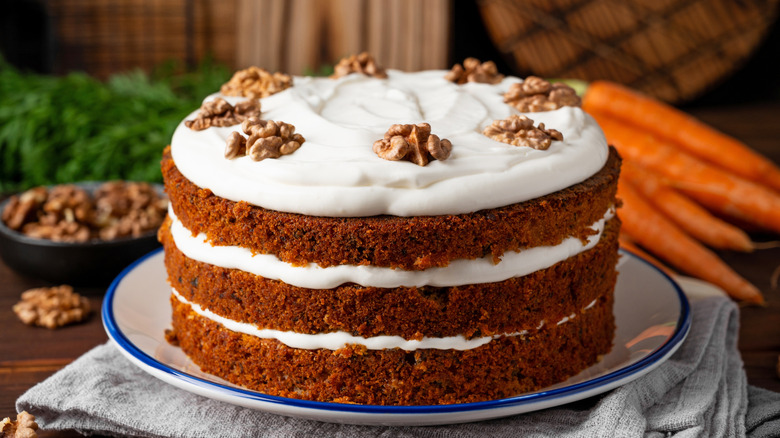 Carrot cake with frosting, nuts, and carrots in background