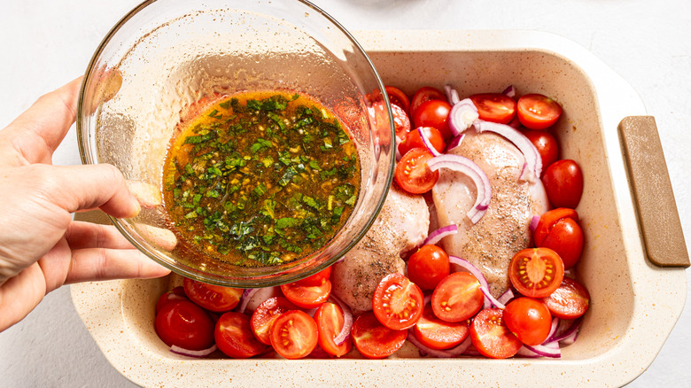 Adding sauce to a baking dish with chicken, cherry tomatoes, and red onion