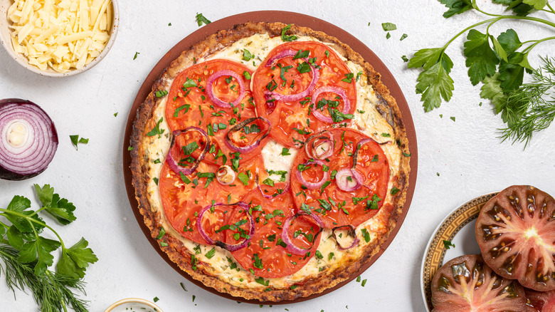 Roasted tomato pie surrounded by slices of fresh tomatoes, fresh parsley, dill, red onion, and shredded cheese