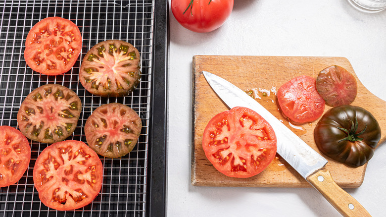 Cutting board with a knife, sliced tomatoes, and more tomato slices on а wired rack
