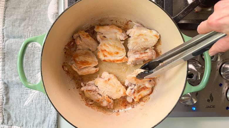 Chicken thighs browning in butter in Dutch oven on stovetop with tongs