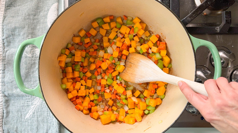 Stirring onion, celery, carrot, and butternut squash cubes with wooden spoon in Dutch oven on stovetop