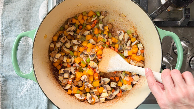 Stirring mushrooms, butternut squash, and mirepoix in Dutch oven with wooden spoon