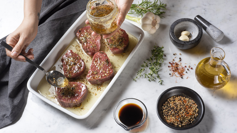 Person spooning marinade over steaks with herbs and other ingredients surrounding the white pan