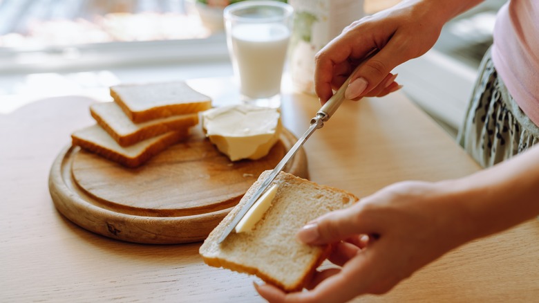 person's hand spreading butter on slice of toasted bread, glass of fresh milk in background