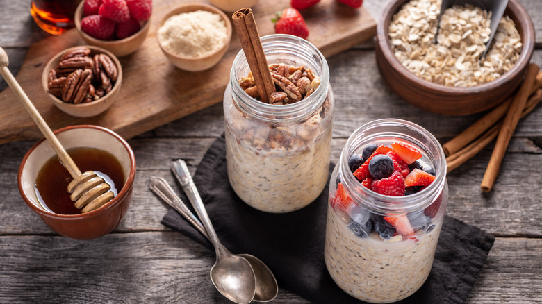 Two jars of overnight oats, one topped with nuts, a cinnamon stick stuck in it, the other topped with fresh mixed berries, two spoons below, surrounded by dry oats, cinnamon sticks, honey, pecans, raspberries, and strawberries