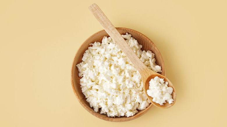 A wooden bowl of cottage cheese with a wooden spoon laid across it, the spoon full of cottage cheese, all on a light brown countertop.