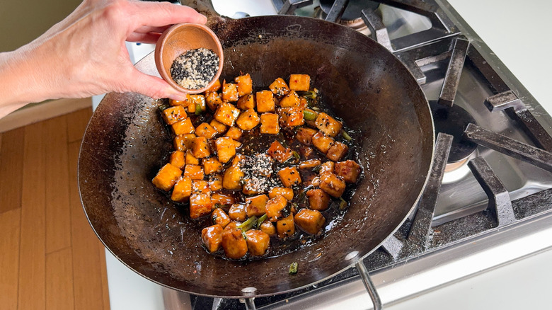 Sprinkling white and black sesame seeds on crispy and sticky baked sesame tofu in wok on stovetop