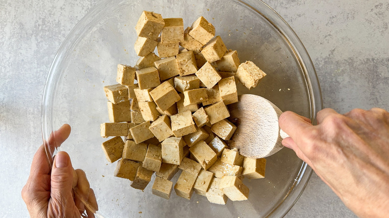 Mixing cubes of tofu with soy sauce and sesame oil in large glass bowl with wooden spoon