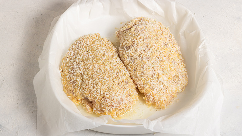 Two chicken breasts covered in coconut batter on a parchment paper lined baking dish