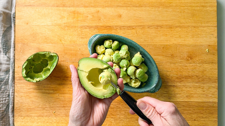 Scooping avocado flesh out of shell with melon baller on cutting board