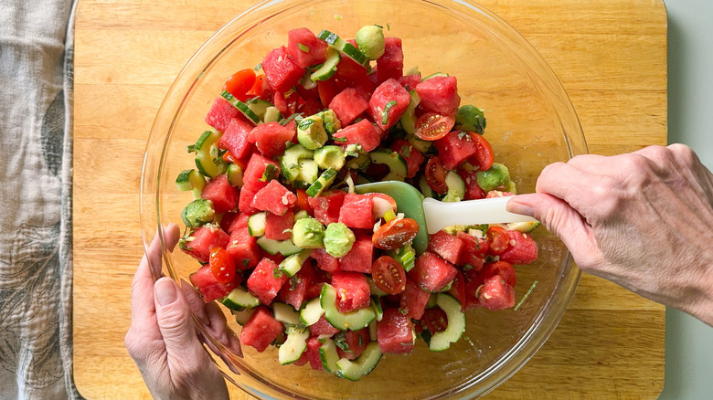 Mixing avocado balls and herbs into deconstructed watermelon salad in glass bowl with rubber spatula
