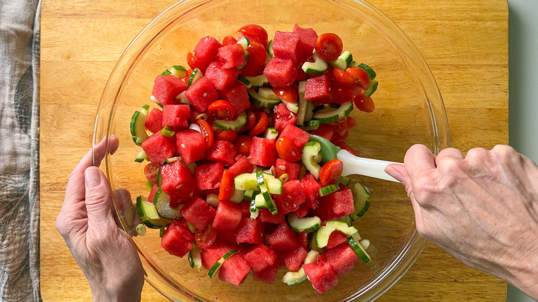 Mixing cut watermelon, cucumber, grape tomatoes, and scallions on glass bowl on cutting board