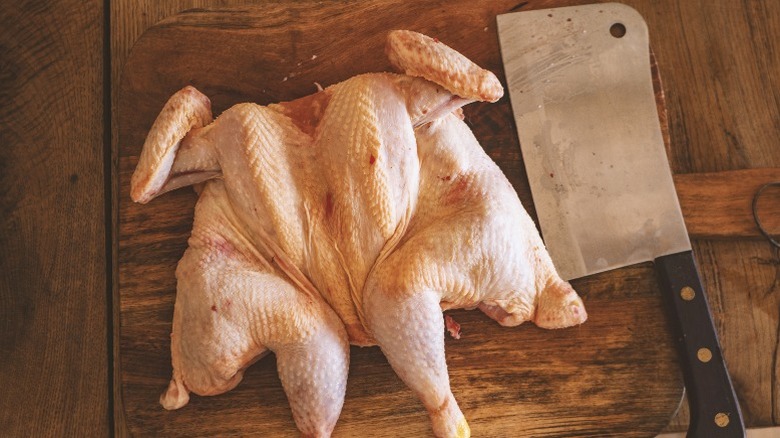 A spatchcocked chicken lies on a cutting board next to a cleaver