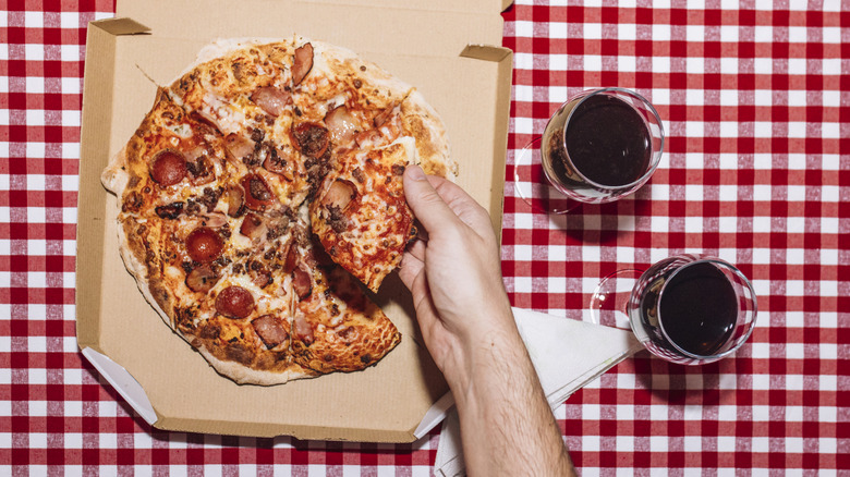 Person picking up a slice of pepperoni pizza next to two glasses of red wine on a checkered background